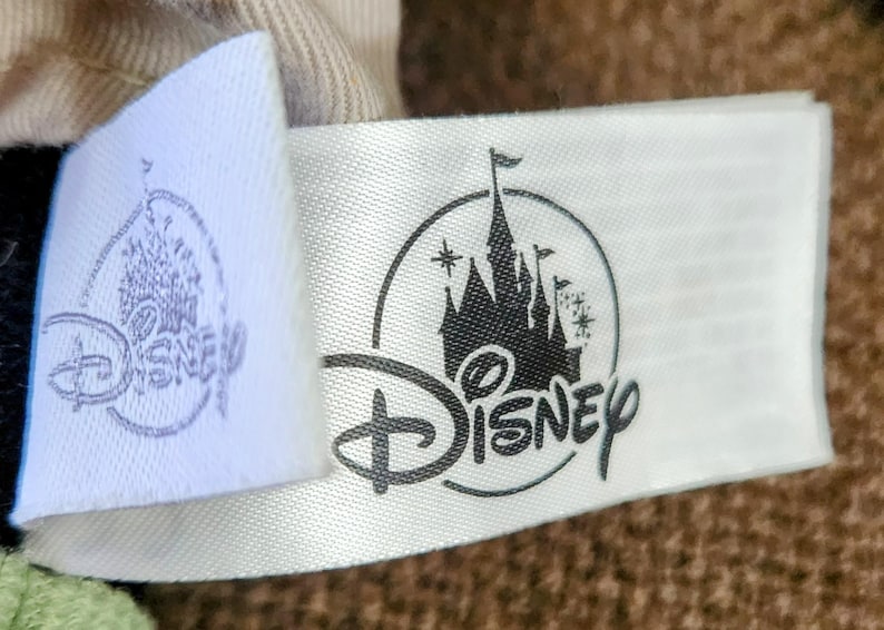 Puede incluir: Primer plano de una etiqueta de tela blanca con el logotipo de Disney y una silueta negra de un castillo. El logotipo de Disney est&aacute; en una fuente cursiva. El castillo tiene m&uacute;ltiples torres y una bandera.