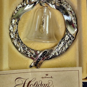 May include: A silver-toned Christmas ornament in the shape of a wreath with a bow at the top and a glass bell in the center. The ornament has a banner with the year "1987" and is labeled "Holiday Heirloom" by Hallmark.