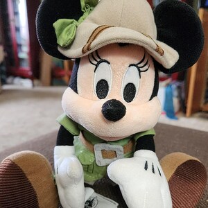 Puede incluir: Mu&ntilde;eca de peluche de Minnie Mouse con un atuendo de safari. La mu&ntilde;eca tiene orejas negras, un sombrero beige con un lazo verde y una camisa verde con un cintur&oacute;n marr&oacute;n. Las manos son blancas y los pies marrones.