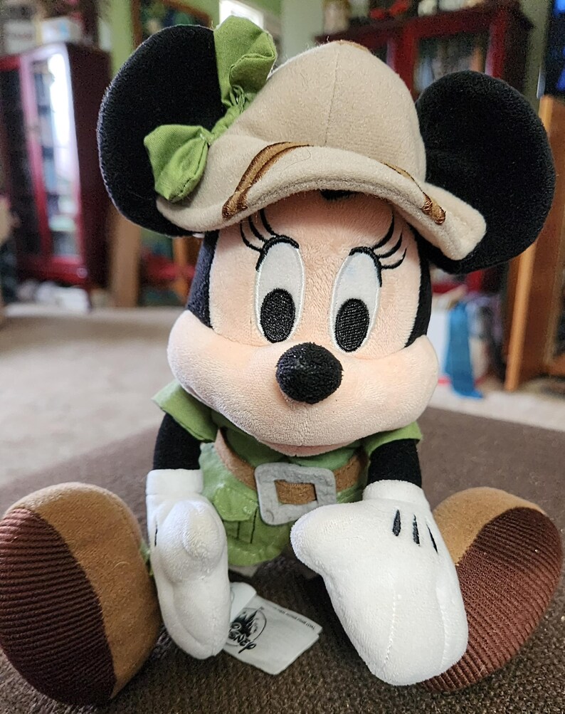 Puede incluir: Mu&ntilde;eca de peluche de Minnie Mouse con un atuendo de safari. La mu&ntilde;eca tiene orejas negras, un sombrero beige con un lazo verde y una camisa verde con un cintur&oacute;n marr&oacute;n. Las manos son blancas y los pies marrones.