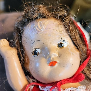 May include: A vintage doll with a painted face, brown hair, and a red hood. The doll has detailed facial features, including painted eyes and a red mouth. The doll is wearing a red dress with white lace trim. The doll's arm is raised.