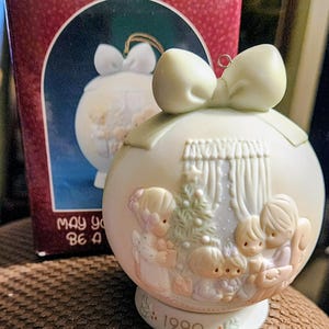 May include: A round, cream-colored porcelain Christmas ornament featuring a sculpted relief of four children gathered around a decorated tree. The ornament is topped with a large bow and has the year 1990 printed on the base, accompanied by its box.