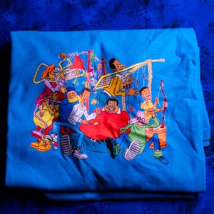 以下が含まれることがあります： 折りたたまれた、明るい青色のTシャツ。カラフルな漫画のイラストには、数人の人々が描かれています。イラストは、楽器を持った人々と弓矢を持った人を示しています。アートワークは鮮やかで詳細です。