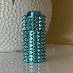 May include: A cylindrical teal container with a textured surface featuring rows of raised, pointed pyramid studs. The object has a flat, smooth circular cap at the top and a matching teal finish throughout, reflecting light on its metallic-like surface.