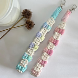 Puede incluir: Dos llaveros de macramé con cierres plateados. Uno es azul pastel, blanco y amarillo, el otro rosa y blanco. Los llaveros tienen un diseño tejido con nudos decorativos, sobre una superficie blanca.