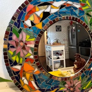 May include: A round wall mirror featuring a decorative mosaic frame composed of multi-colored glass tiles. The frame displays a vibrant pattern of pink floral motifs, bright orange shapes, and various shades of blue, green, and yellow glass pieces.