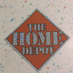 May include: An orange diamond-shaped sticker with the text "THE HOME DEPOT" in silver glitter lettering. The diamond is outlined with a silver glitter border. The sticker is on a speckled white surface.