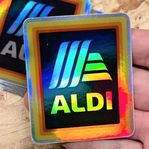 May include: A holographic sticker with a rainbow border featuring the ALDI logo. The logo is a stylized letter A with three stripes and the word ALDI in yellow letters.