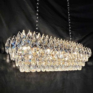 May include: A rectangular chandelier with numerous clear, teardrop-shaped crystals. The crystals are arranged in multiple layers, creating a sparkling effect. The fixture hangs from two silver chains against a dark background.