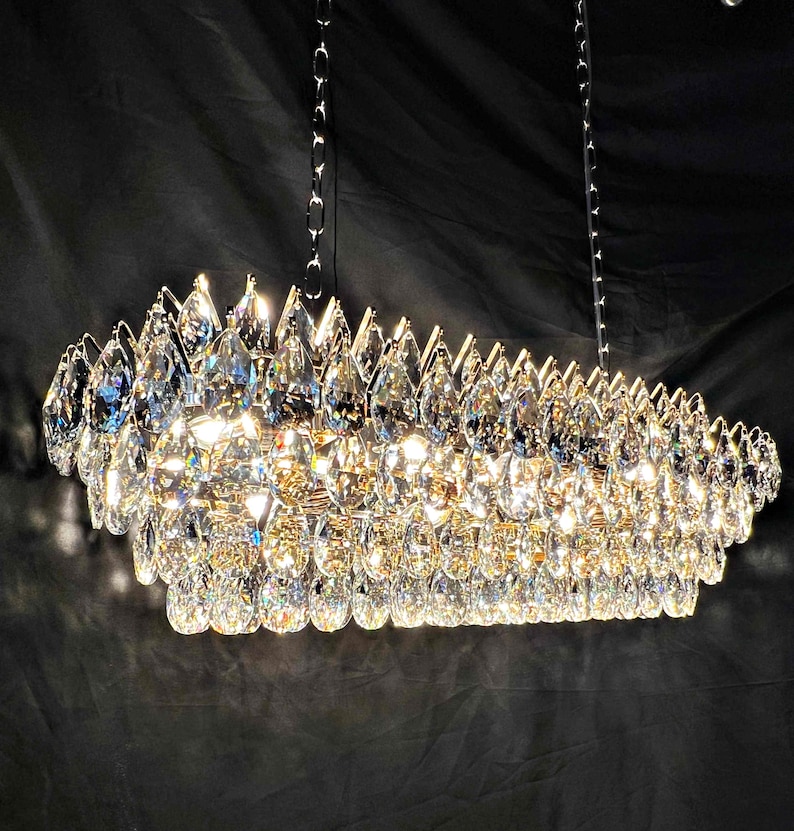 May include: A rectangular chandelier with numerous clear, teardrop-shaped crystals. The crystals are arranged in multiple layers, creating a sparkling effect. The fixture hangs from two silver chains against a dark background.
