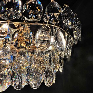 May include: Close-up of a crystal chandelier with multiple teardrop-shaped crystals. The crystals are clear and faceted, reflecting light. The chandelier has a gold-colored metal frame. The background is black.