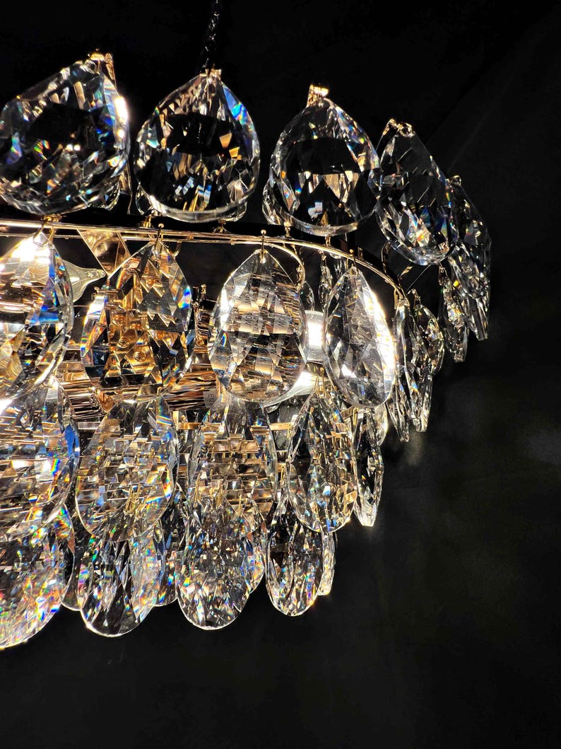 May include: Close-up of a crystal chandelier with multiple teardrop-shaped crystals. The crystals are clear and faceted, reflecting light. The chandelier has a gold-colored metal frame. The background is black.