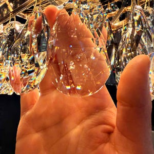 May include: Close-up of a crystal chandelier with multiple clear, faceted crystal pendants. The crystals are suspended from a gold-colored metal frame. The light refracts through the crystals, creating a rainbow effect.