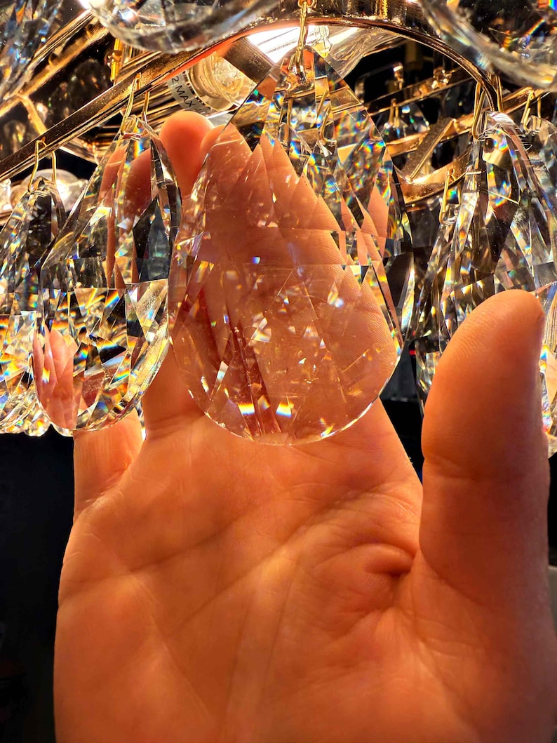 May include: Close-up of a crystal chandelier with multiple clear, faceted crystal pendants. The crystals are suspended from a gold-colored metal frame. The light refracts through the crystals, creating a rainbow effect.