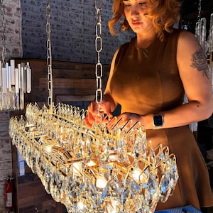 May include: A rectangular gold chandelier with multiple crystal pendants. The chandelier is suspended from a chain. The image shows a woman in a brown dress working on the chandelier.