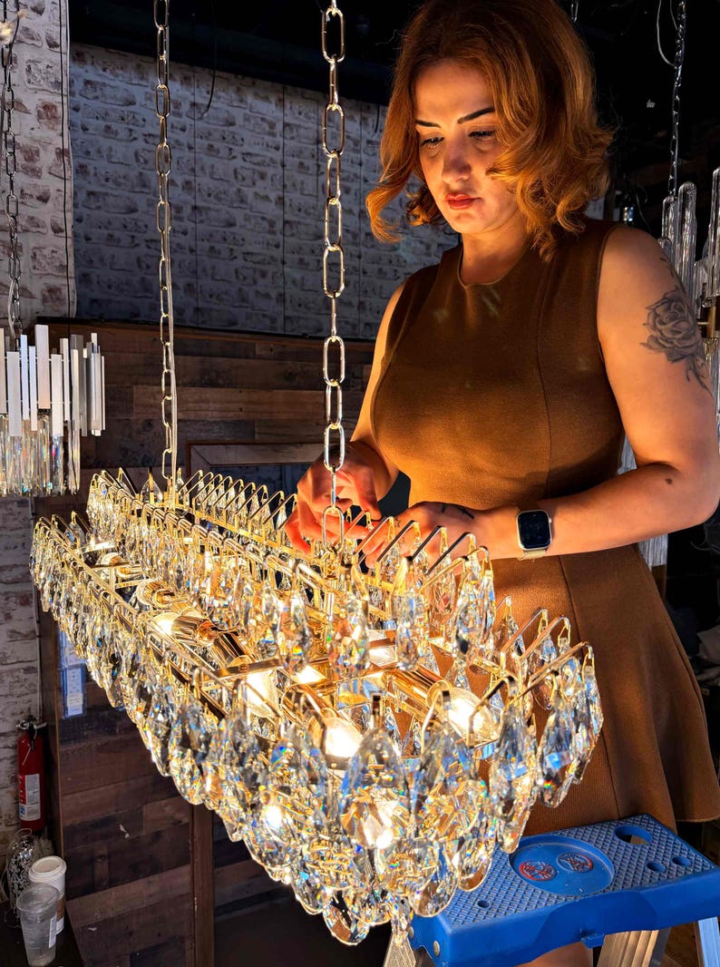 May include: A rectangular gold chandelier with multiple crystal pendants. The chandelier is suspended from a chain. The image shows a woman in a brown dress working on the chandelier.