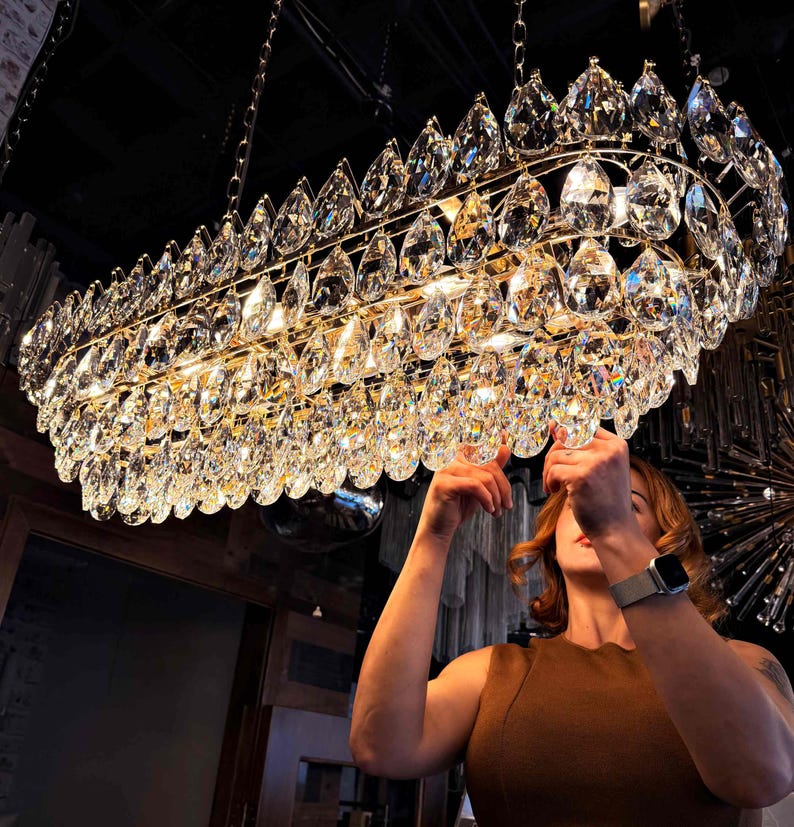 May include: A large, rectangular chandelier with multiple tiers of clear, teardrop-shaped crystals. The crystals are suspended from a gold-colored metal frame. The chandelier is hanging from the ceiling.