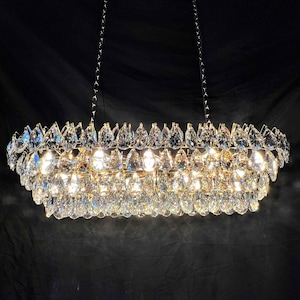 May include: A rectangular chandelier with multiple tiers of clear, teardrop-shaped crystals. The fixture is suspended by chains and emits a bright, warm light, creating a sparkling effect against a dark background. A luxurious lighting fixture.
