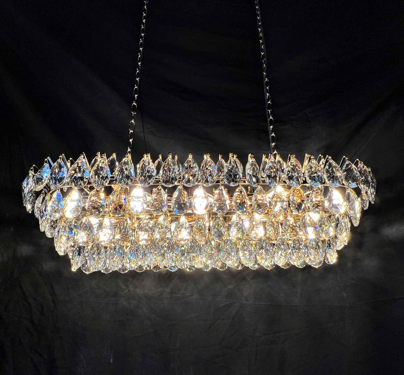 May include: A rectangular chandelier with multiple tiers of clear, teardrop-shaped crystals. The fixture is suspended by chains and emits a bright, warm light, creating a sparkling effect against a dark background. A luxurious lighting fixture.