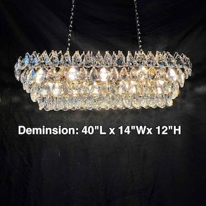 May include: A rectangular crystal chandelier with multiple tiers of sparkling crystals. The dimensions are 40"L x 14"W x 12"H. The fixture hangs from two chains and is illuminated, creating a dazzling display of light and reflection.