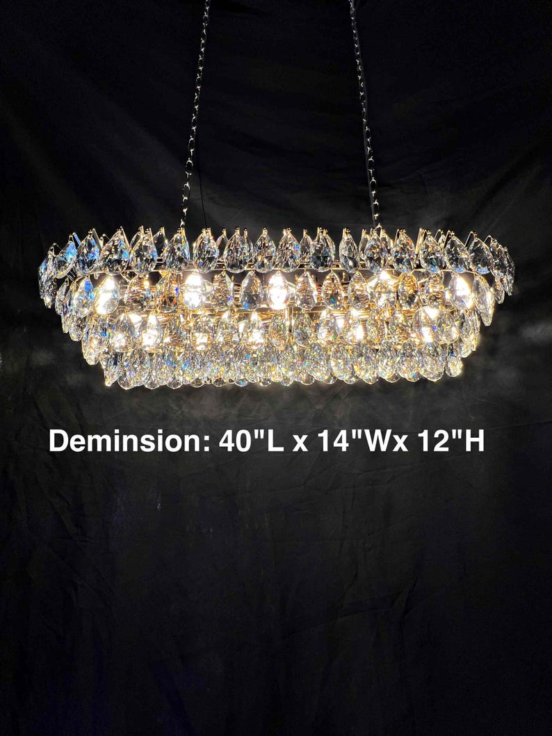 May include: A rectangular crystal chandelier with multiple tiers of sparkling crystals. The dimensions are 40"L x 14"W x 12"H. The fixture hangs from two chains and is illuminated, creating a dazzling display of light and reflection.