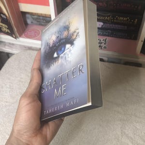 May include: A paperback book titled "Shatter Me" by Tahereh Mafi. The cover features a blue eye with a tree-like design and the title in silver lettering. The book is held in a person's hand, with other books visible in the background.