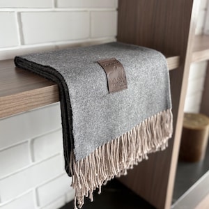Puede incluir: Una manta de lana de alpaca gris doblada con un borde con flecos. Una etiqueta de cuero marrón con las palabras "Alpaca Glory" y un logotipo está adherida a la manta. La manta se exhibe en una estantería de madera.