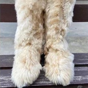 Könnte beinhalten: Ein Paar cremefarbene, flauschige Stiefel. Die Stiefel scheinen aus einem weichen, strukturierten Material zu bestehen, möglicherweise aus Kunstfell oder einem ähnlichen Stoff. Die Stiefel sind kniehoch und haben eine runde Spitze.