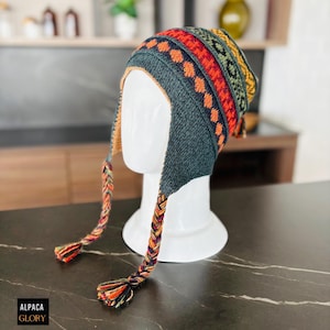 Puede incluir: Un gorro de punto gris con orejeras y un colorido estampado geométrico. El gorro presenta rayas naranjas, rojas, verdes y amarillas. Borlas trenzadas cuelgan de las orejeras. El gorro se exhibe en una cabeza de maniquí blanca.