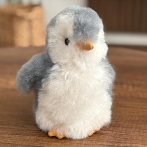 May include: A plush penguin toy with a soft, fluffy texture. The penguin has a gray head and wings, a white body, and a small, orange beak and feet. The toy is designed to resemble a baby penguin.