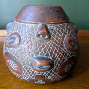 May include: A round, brown, carved vessel featuring a textured cross-hatched pattern across its surface. The object displays raised, sculpted facial features including a nose and mouth, integrated into the overall design of the container.