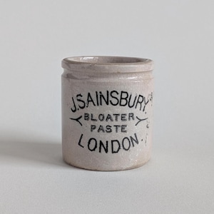 May include: A cylindrical ceramic pot with a light beige, crackled glaze finish. The front features black printed text that reads J.SAINSBURY BLOATER PASTE LONDON, with decorative arrow-like symbols flanking the word BLOATER.