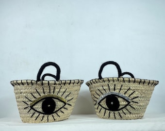 Puede incluir: Dos bolsas de mano de paja con asas negras y diseños de ojos. Un ojo tiene purpurina dorada y el otro tiene purpurina plateada.