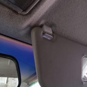 Puede incluir: Un parasol de coche gris claro está parcialmente abierto, revelando un vistazo del techo interior y una parte del espejo retrovisor. El parasol está sujeto al techo con un clip blanco.