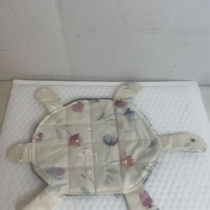 May include: A beige quilted turtle-shaped item with a floral pattern in shades of red, blue, and purple. The turtle has four flipper-like appendages and a dark border. The item is likely a decorative piece or a kitchen accessory.