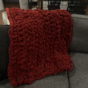May include: A red knitted throw blanket with a chunky knit texture. The blanket is draped over a gray couch.