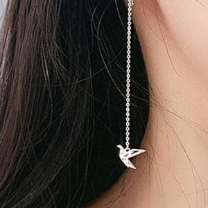 Può includere: Orecchino in argento con una delicata catena e un piccolo ciondolo a forma di uccello. L'orecchino è progettato per pendere dall'orecchio, creando un look semplice ma elegante. Il ciondolo a forma di uccello aggiunge un tocco di fantasia.