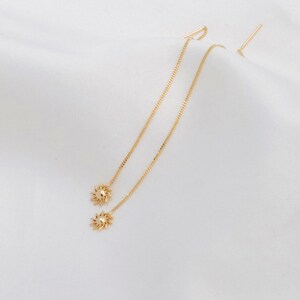 Può includere: Orecchini pendenti color oro con un motivo a forma di sole all'estremità di ogni catena. Gli orecchini hanno una delicata catena che pende dall'orecchio. Gli orecchini sono esposti su sfondo bianco.