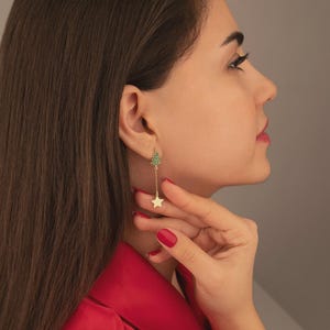 Può includere: Un orecchino dorato con un design ad albero di Natale adornato con pietre preziose verdi e una stella pendente. L'orecchino è indossato da una persona che indossa una giacca rossa. Lo sfondo è grigio neutro.