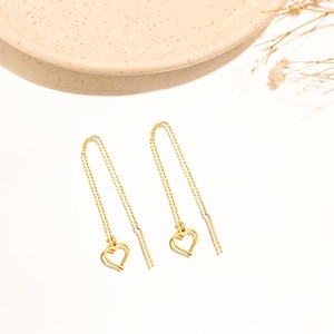 Può includere: Un paio di orecchini pendenti color oro. Ogni orecchino presenta una delicata catena che conduce a un ciondolo a forma di cuore e una barretta dritta. Presentati su una superficie bianca.