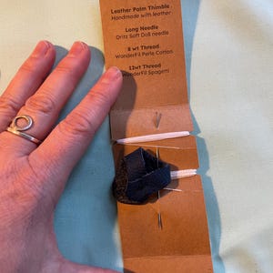 May include: A brown paper package containing a hand sewing kit. The kit includes a leather palm thimble, a long needle, 8wt thread, and 12wt thread. The package is labelled "Hand Sewing Kit".