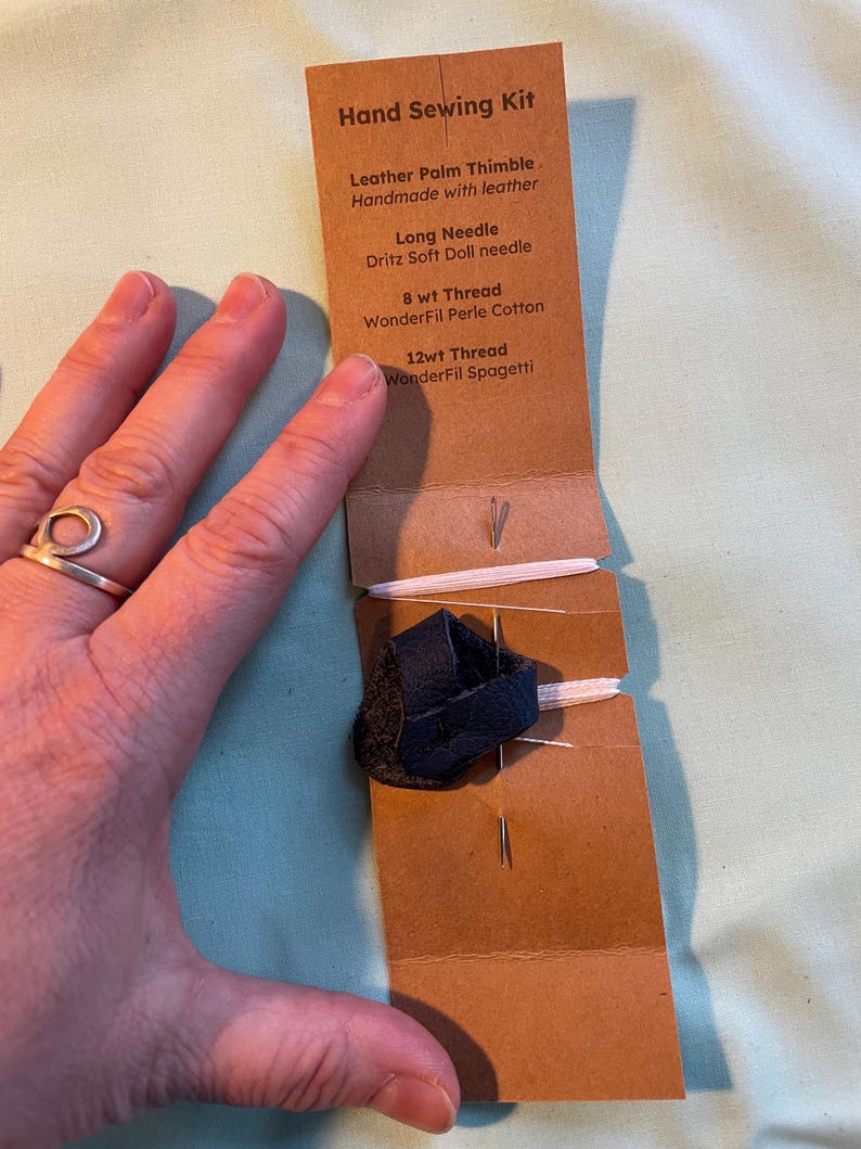 May include: A brown paper package containing a hand sewing kit. The kit includes a leather palm thimble, a long needle, 8wt thread, and 12wt thread. The package is labelled "Hand Sewing Kit".
