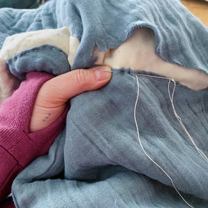 May include: A close-up of a hand stitching two pieces of blue fabric together with white thread. The fabric is a light blue colour and has a soft, textured feel.