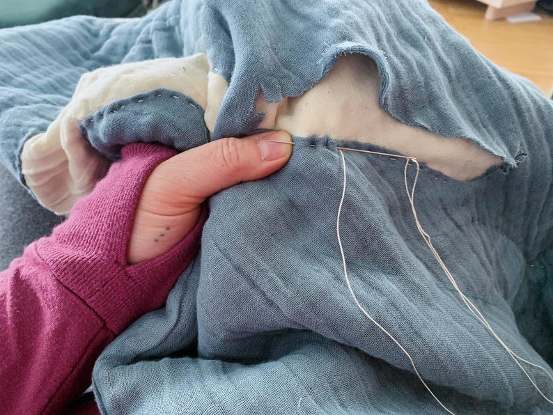 May include: A close-up of a hand stitching two pieces of blue fabric together with white thread. The fabric is a light blue colour and has a soft, textured feel.