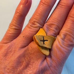 May include: A tan leather ring with a cross stitched on it. The ring is worn on a finger.