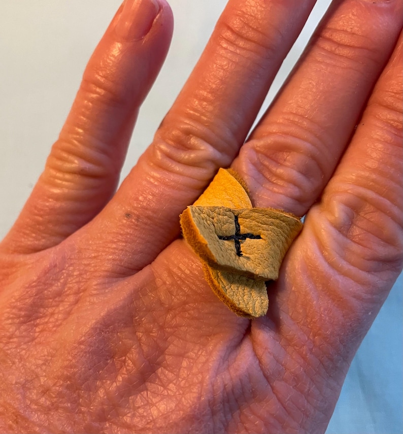 May include: A tan leather ring with a cross stitched on it. The ring is worn on a finger.