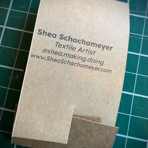 May include: A brown business card with the text "Shea Schachameyer Textile Artist @shea.making.doing www.SheaSchachameyer.com" printed in black.