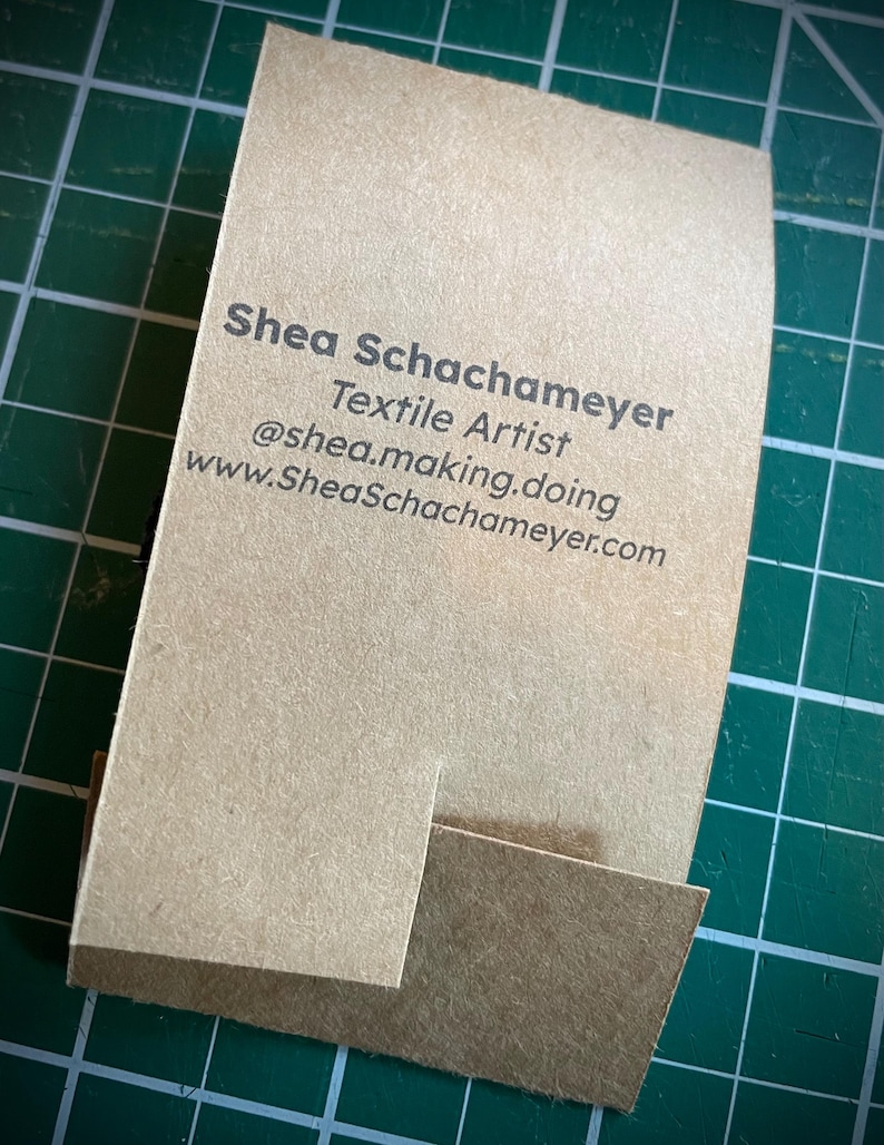 May include: A brown business card with the text "Shea Schachameyer Textile Artist @shea.making.doing www.SheaSchachameyer.com" printed in black.