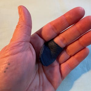 May include: A close-up of a hand wearing a dark blue leather fingertip protector. The protector is worn on the index finger and is made of soft, pliable leather.