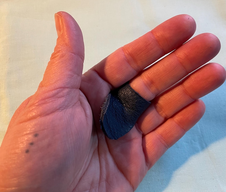 May include: A close-up of a hand wearing a dark blue leather fingertip protector. The protector is worn on the index finger and is made of soft, pliable leather.